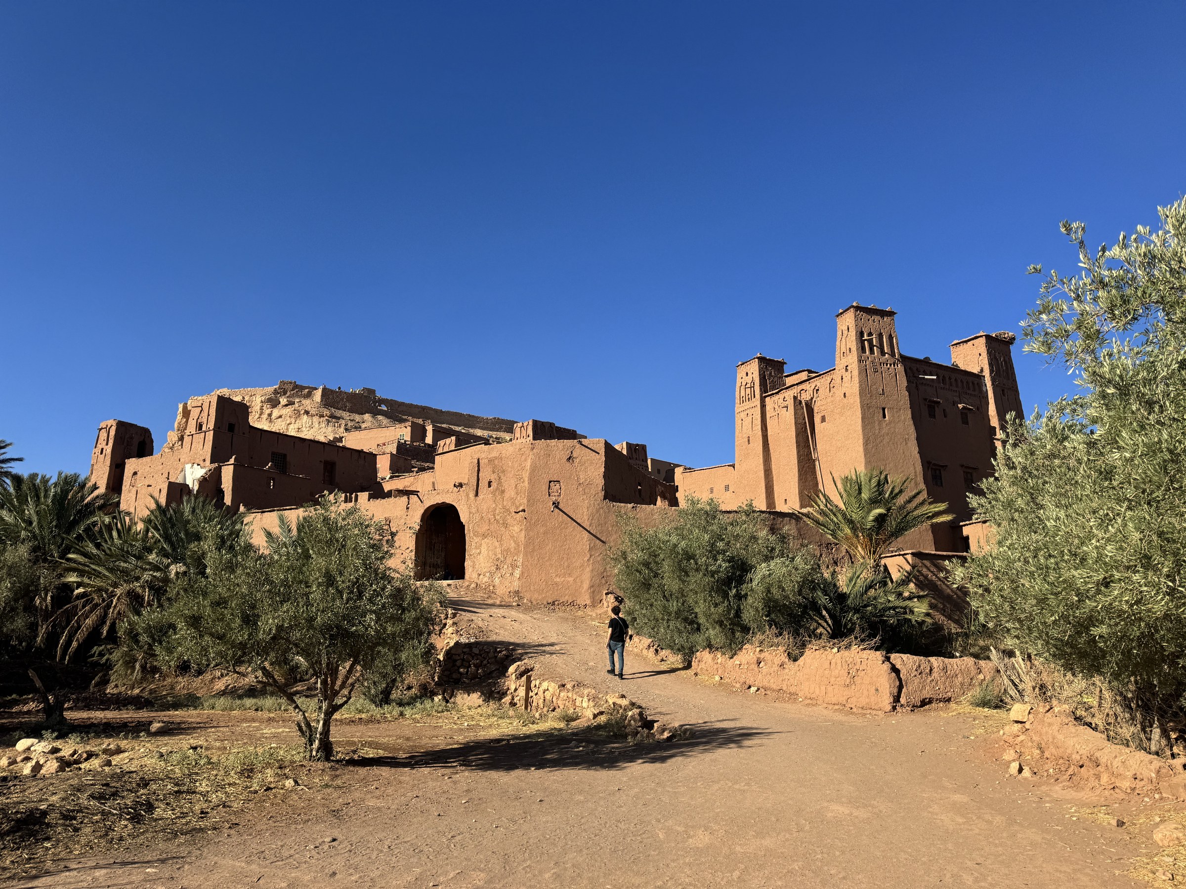 Kasbah de Ait Ben Haddou desde el camino de acceso, torres de barro rojizo y palmeras bajo cielo azul, Marruecos