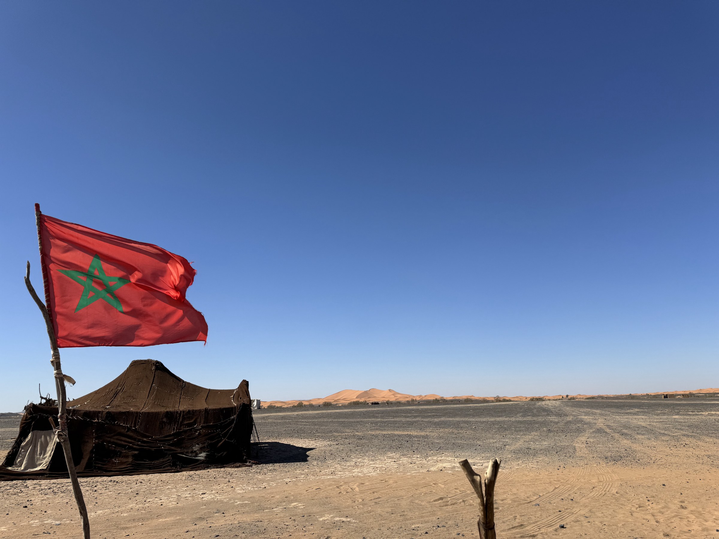 Bandera de Marruecos ondeando junto a una jaima n&oacute;mada con las dunas del Erg Chebbi al fondo, desierto de Merzouga