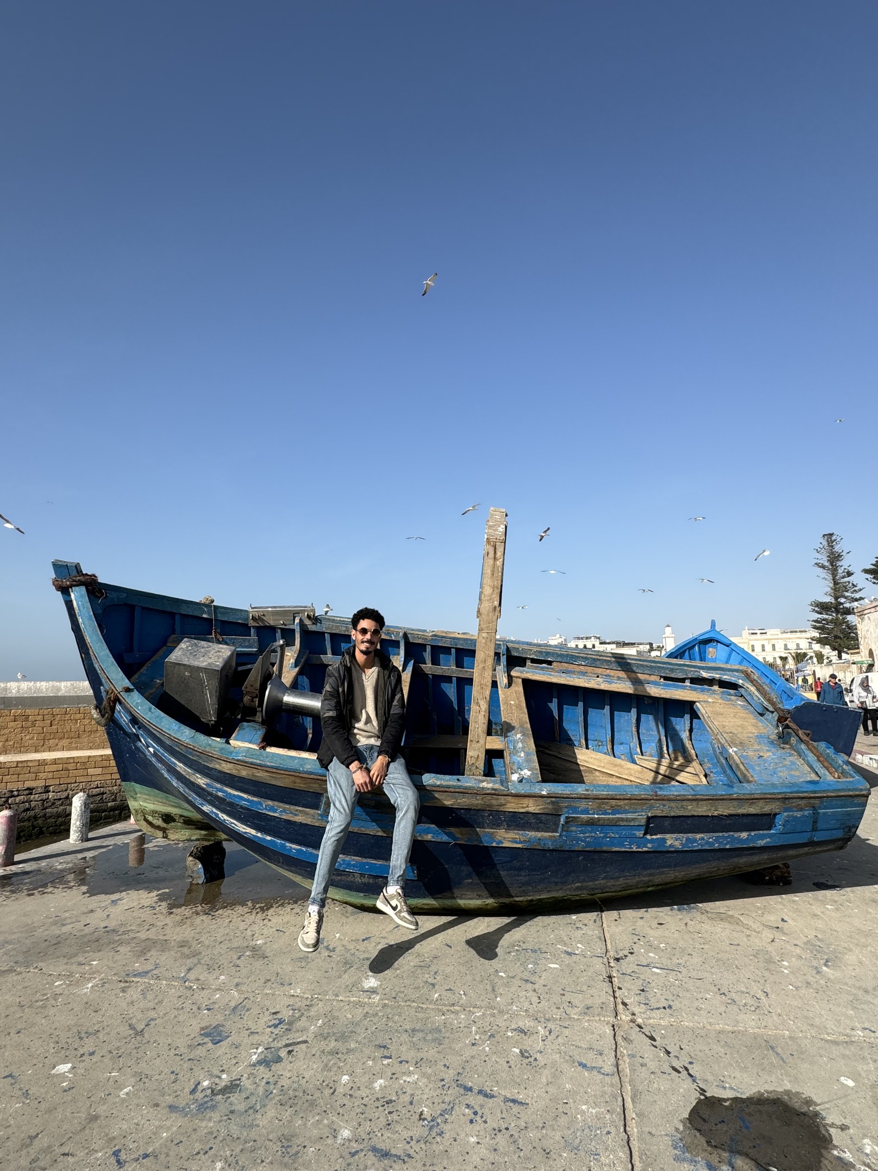 Hombre apoyado en barca de pesca azul tradicional en el puerto de Essaouira con gaviotas volando